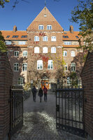 An old building at LUX against a blue sky. The leaves on the building have turned red. Three people wearing big coats are walking towards the building.