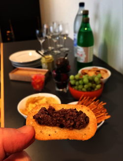 A white hand holds a cracker with mushroom cream on it. In the background, a spread of snacks and soft drinks.