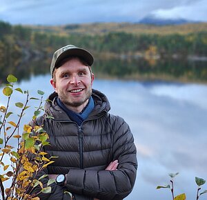 Kirill Elin is a white person dressed in a puffer jacket. He is smiling into the camera with his arms crossed. The photo is taken in front of a lake. In the far background, a forest in front of a mountain.