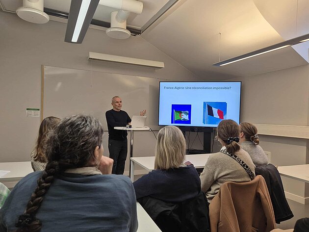 Thierry Gilles is speaking next to a presentation projected on a screen. The audience is seated and seen from behind.