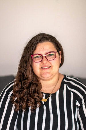 Henriette is a white person with long curly hair. She is wearing a black and white top and red glasses. She is looking into the camera and smiling.