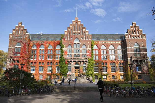 A photo of the university against a blue sky. The building is covered in green, yellow and red leaves.