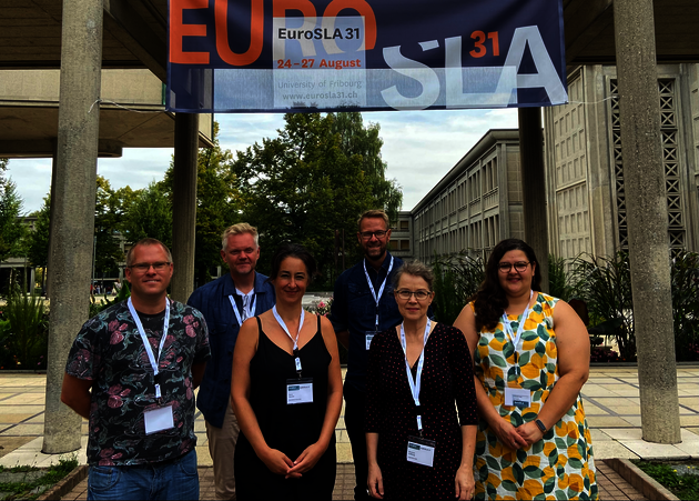 Six LAMiNATE members stand in front of a banner that reads "EuroSLA 31." The sun is shining. Some of the people are smiling, some are squinting.